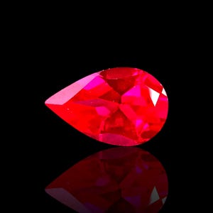 May include: A faceted, pear-shaped, bright red gemstone against a black background. The gem has multiple facets that reflect light, creating a sparkling effect. The gem's reflection is visible below it.