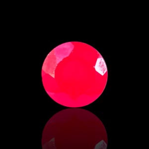 Op de afbeelding: Een gefacetteerde, ronde, helderrode edelsteen tegen een zwarte achtergrond. De edelsteen heeft een reflecterend oppervlak en de weerspiegeling ervan is hieronder zichtbaar. De facetten van de edelsteen vangen het licht op en creëren highlights en schaduwen.