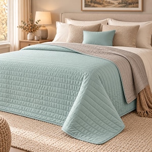 May include: A light blue quilted bedspread with a linear stitched pattern draped over a bed. The bedding set includes two large beige textured pillows, two white pillows, and a small rectangular light blue accent pillow placed on top.