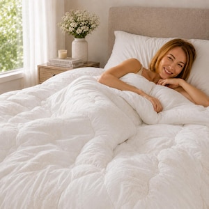 Peut inclure: Une couette blanche moelleuse avec une texture matelassée recouvrant un lit. La literie présente une surface douce et touffetée drapée sur un matelas. Le tissu blanc possède un motif gaufré répétitif subtil sur toute sa surface.