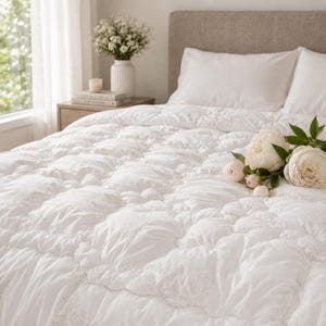 May include: A white quilted comforter with a textured, puffed surface pattern covering a bed. The bedding features a soft, plush appearance with a grid-like stitching design. A bouquet of white peony flowers rests on the surface of the comforter.