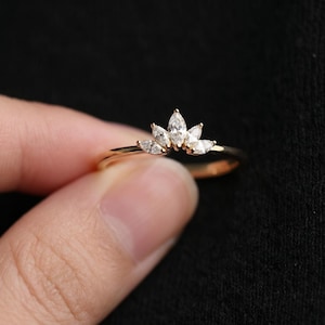 May include: A gold-colored ring featuring a crown design with marquise-cut clear gemstones. The ring's band is thin and delicate, and the gemstones are arranged in a curved pattern, creating a sparkling effect. The ring is shown against a dark background.