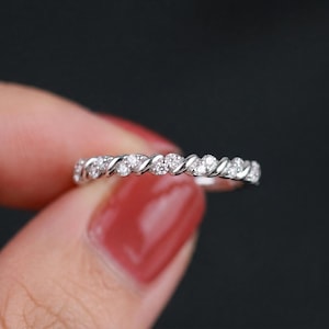 Peut inclure: Une bague en argent avec un motif de bande torsadée, ornée de petites pierres blanches scintillantes. La bague est présentée sur un fond sombre, mettant en valeur ses détails complexes et son style élégant.