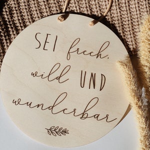 May include: A round wooden sign with the text "Sei frech, wild und wunderbar" (Be bold, wild and wonderful) in brown lettering. A small leaf design is below the text.