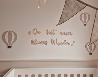 May include: A white crib with a white rope bumper and a light brown wall with a wooden sign that says "Du bist unser kleines Wunder" with a heart on each side of the text. There are three hot air balloon decorations on the wall, two are light brown and one is gray.