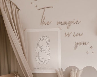 Può includere: Una cameretta per bambini con una culla di legno, una parete bianca con le parole "The magic is in you" in lettere di legno, una stampa di un bambino in posizione fetale e un vaso con erba della pampa secca.