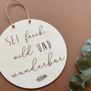 May include: A round wooden sign with a brown rope hanger. The sign has the text "Sei frech, wild und wunderbar" written in brown cursive lettering with a small leaf design at the bottom.