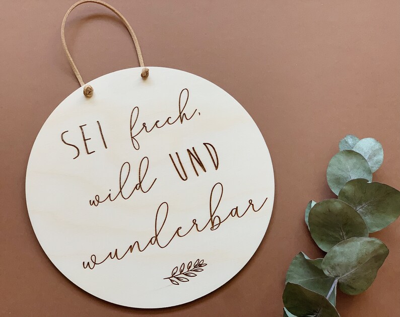 May include: A round wooden sign with a brown rope hanger. The sign has the text "Sei frech, wild und wunderbar" written in brown cursive lettering with a small leaf design at the bottom.