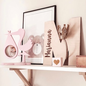 Pu&ograve; includere: Una cicogna di legno rosa con una cornice per foto per bambini, un'immagine di un orso di legno, una lettera M di legno con il nome "Marianna" inciso su di essa, una corona dorata e un blocco di cuore di legno su uno scaffale bianco.