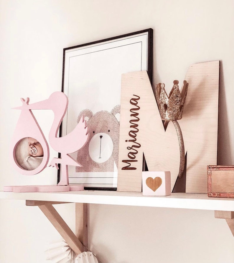 Pu&ograve; includere: Una cicogna di legno rosa con una cornice per foto per bambini, un'immagine di un orso di legno, una lettera M di legno con il nome "Marianna" inciso su di essa, una corona dorata e un blocco di cuore di legno su uno scaffale bianco.