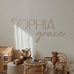Op de afbeelding: Een lichtbruin houten bord met de naam "Sophia grace" in cursief schrift. Het bord hangt aan een witte muur in een kinderkamer. Er is een klein houten poppenhuis, een rieten tafel en stoelen en knuffels in de kamer.