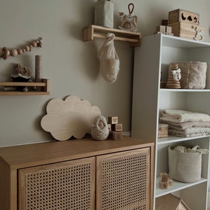 May include: A light wood cabinet with two doors featuring a woven rattan design. The cabinet is on a wall with a white shelf and a wooden cloud-shaped shelf. The shelf has a small knitted animal and wooden blocks with letters and symbols.