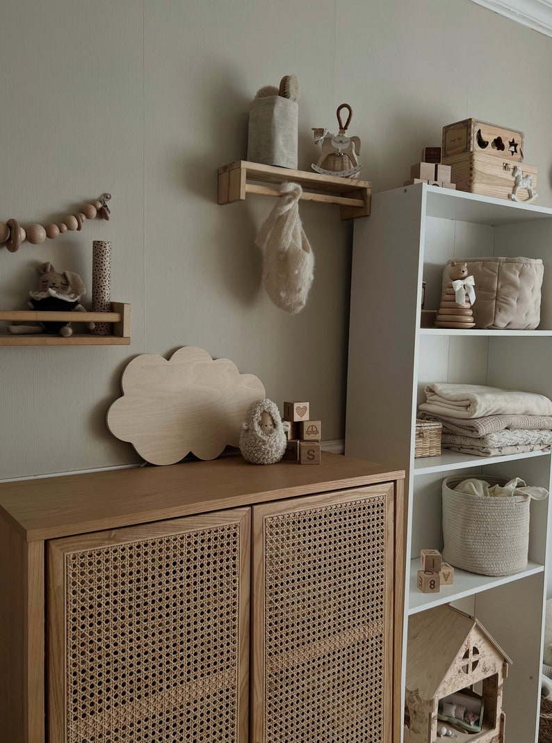 May include: A light wood cabinet with two doors featuring a woven rattan design. The cabinet is on a wall with a white shelf and a wooden cloud-shaped shelf. The shelf has a small knitted animal and wooden blocks with letters and symbols.