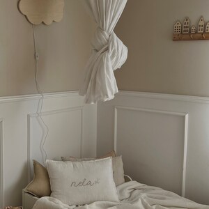 May include: A white canopy bed with a white blanket and a pillow with the word "rela" embroidered on it. A wooden cloud nightlight is on the wall above the bed.