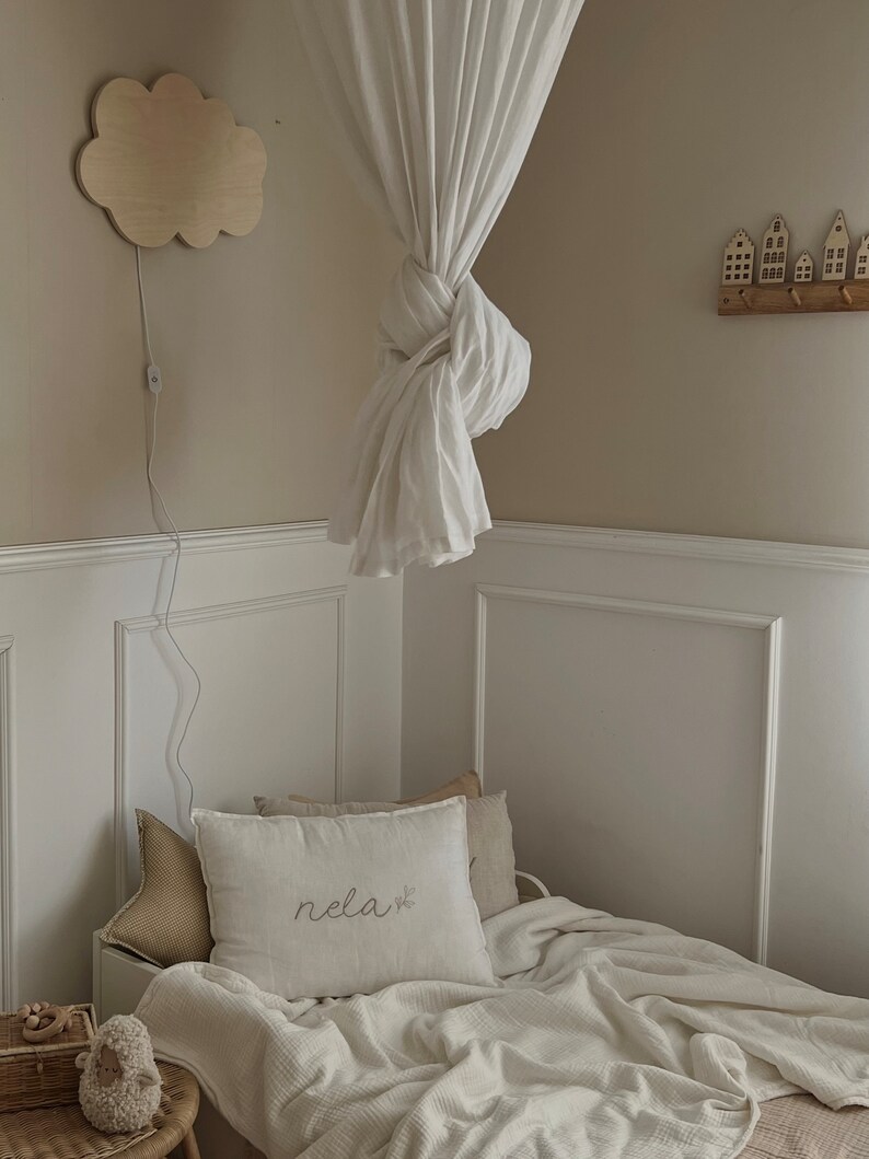 May include: A white canopy bed with a white blanket and a pillow with the word "rela" embroidered on it. A wooden cloud nightlight is on the wall above the bed.
