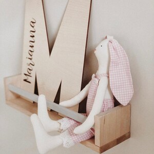 Pu&ograve; includere: Uno scaffale in legno con una barra di metallo grigia, una lettera in legno 'M' con il nome "Marianna" inciso su di essa e un coniglio di peluche rosa e bianco a quadri seduto sullo scaffale.