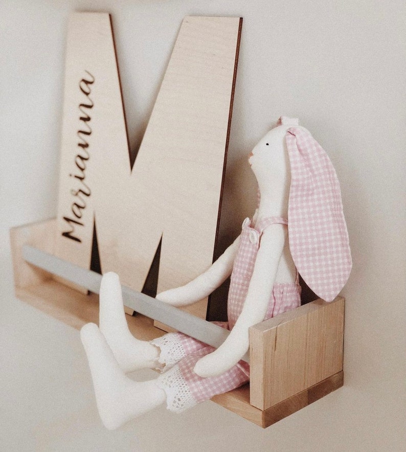Pu&ograve; includere: Uno scaffale in legno con una barra di metallo grigia, una lettera in legno 'M' con il nome "Marianna" inciso su di essa e un coniglio di peluche rosa e bianco a quadri seduto sullo scaffale.