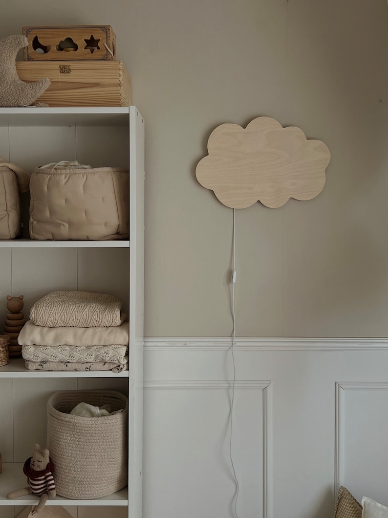 May include: A white bookshelf with a wooden cloud-shaped night light on the wall behind it. The bookshelf has three shelves and is filled with various items, including a wooden toy, a basket, and blankets.