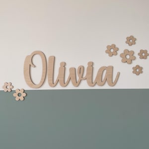 Könnte beinhalten: Holzname "Olivia" in Schreibschrift, mit mehreren Holzblumen-Ausschnitten. Der Name ist an einer zweifarbigen Wand angebracht, mit einem weißen oberen Bereich und einem teal-farbenen unteren Bereich. Dekorative Wandkunst.