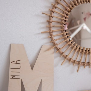 Pu&ograve; includere: Una lettera M in legno con il nome "Mila" inciso su di essa. La lettera &egrave; di colore marrone chiaro e si trova su una superficie bianca. Uno specchio rotondo intrecciato di grandi dimensioni &egrave; sullo sfondo.