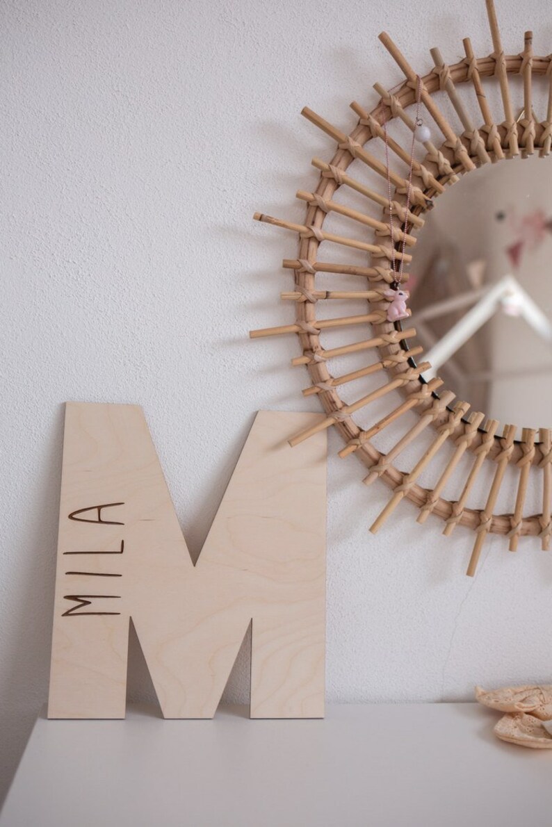 Pu&ograve; includere: Una lettera M in legno con il nome "Mila" inciso su di essa. La lettera &egrave; di colore marrone chiaro e si trova su una superficie bianca. Uno specchio rotondo intrecciato di grandi dimensioni &egrave; sullo sfondo.