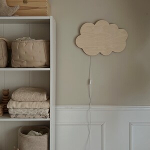 May include: A white wooden shelf unit with three shelves. The top shelf has a wooden toy box with a star, moon, and cloud design. The middle shelf has two beige storage baskets. The bottom shelf has a beige woven basket, a stuffed animal, and a stack of white and beige blankets. A wooden cloud-shaped night light with a white cord is mounted on the wall to the right of the shelf.