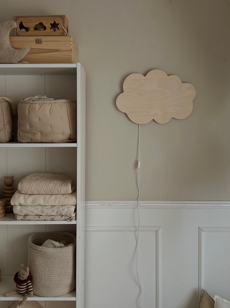 May include: A white wooden shelf unit with three shelves. The top shelf has a wooden toy box with a star, moon, and cloud design. The middle shelf has two beige storage baskets. The bottom shelf has a beige woven basket, a stuffed animal, and a stack of white and beige blankets. A wooden cloud-shaped night light with a white cord is mounted on the wall to the right of the shelf.