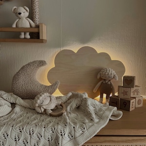 May include: A wooden cloud-shaped night light with a warm glow illuminates a nursery with a crocheted blanket, a stuffed sheep, and wooden blocks with numbers and symbols.