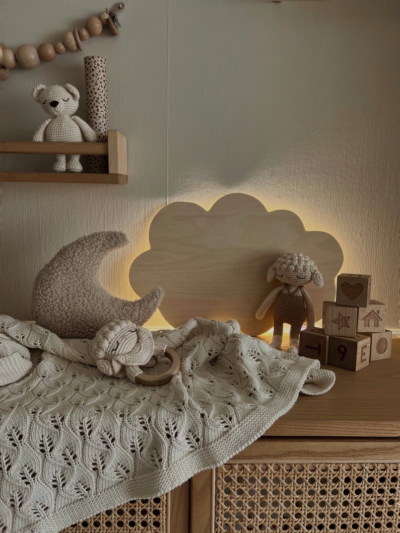 May include: A wooden cloud-shaped night light with a warm glow illuminates a nursery with a crocheted blanket, a stuffed sheep, and wooden blocks with numbers and symbols.