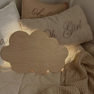 May include: A wooden cloud-shaped night light with a warm glow, sitting on a bed with beige pillows and a blanket. The pillows have the text "Oh Girl" embroidered on them.