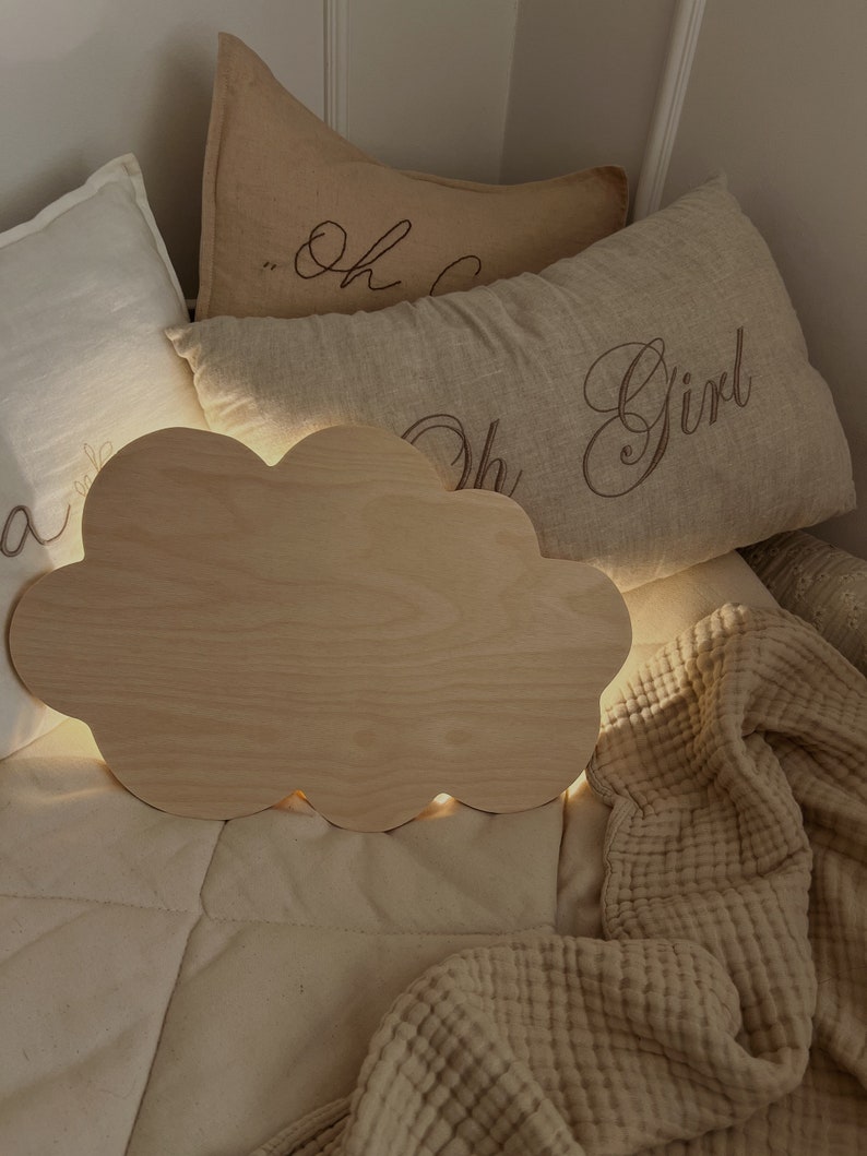 May include: A wooden cloud-shaped night light with a warm glow, sitting on a bed with beige pillows and a blanket. The pillows have the text "Oh Girl" embroidered on them.