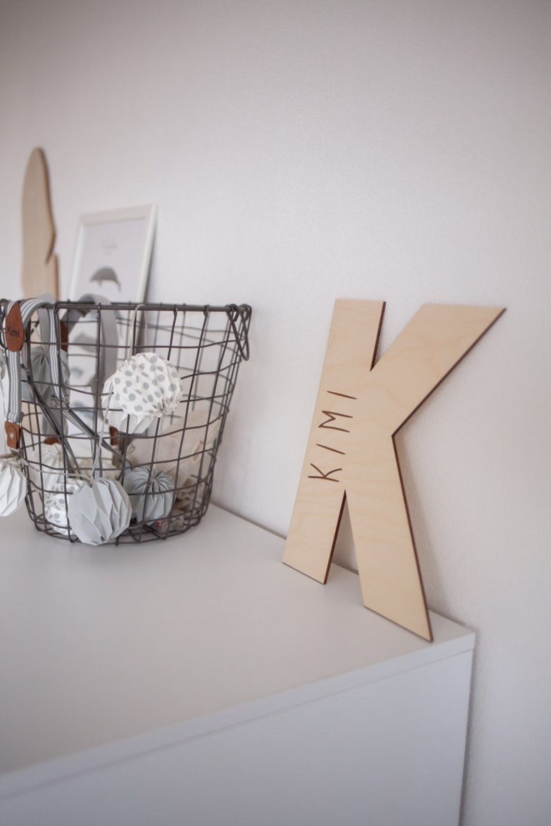 Pu&ograve; includere: Una lettera K in legno con il nome "KIMI" inciso su di essa. La lettera &egrave; appoggiata su una superficie bianca accanto a un cestino di filo metallico pieno di palline di carta bianche e grigie.