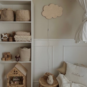May include: A white bookshelf with various items, including baskets, toys, and a dollhouse. A wooden cloud-shaped nightlight is mounted on the wall. A white bed with a white blanket and a pillow with the word "rela" embroidered on it is in the background.