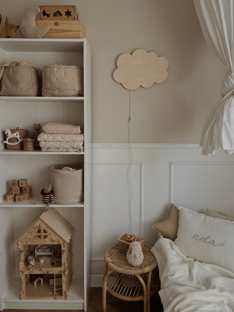 May include: A white bookshelf with various items, including baskets, toys, and a dollhouse. A wooden cloud-shaped nightlight is mounted on the wall. A white bed with a white blanket and a pillow with the word "rela" embroidered on it is in the background.