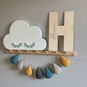 Pu&ograve; includere: Mensola a muro in legno con una mensola a forma di nuvola e una grande lettera H in legno con il nome "Henio" inciso su di essa. La mensola ha una fila di pioli in legno e una ghirlanda di gocce di pioggia in tessuto che pendono sotto.