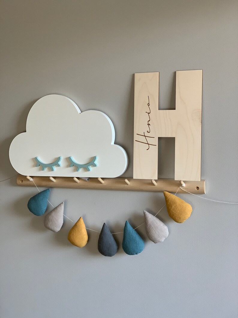 Pu&ograve; includere: Mensola a muro in legno con una mensola a forma di nuvola e una grande lettera H in legno con il nome "Henio" inciso su di essa. La mensola ha una fila di pioli in legno e una ghirlanda di gocce di pioggia in tessuto che pendono sotto.