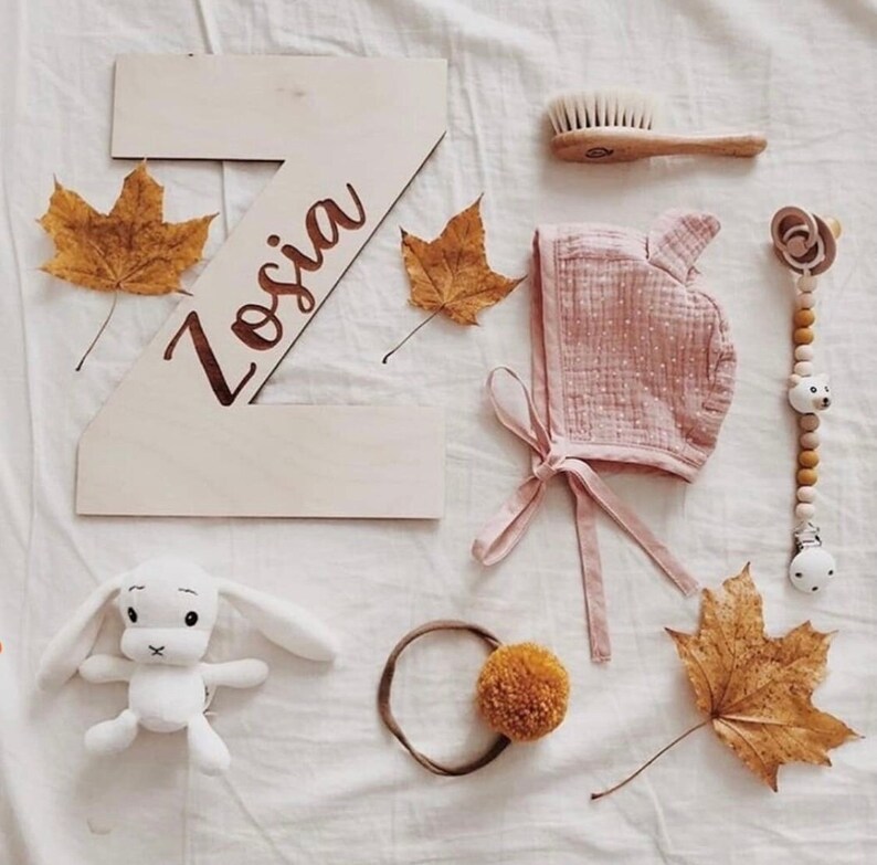 Pu&ograve; includere: Una lettera Z in legno con il nome "Zosia" inciso su di essa, circondata da foglie autunnali, un berretto rosa per neonati, un giocattolo di coniglio bianco, un mordedor di legno, un cerchietto per capelli con un pon pon giallo e una spazzola per capelli in legno.