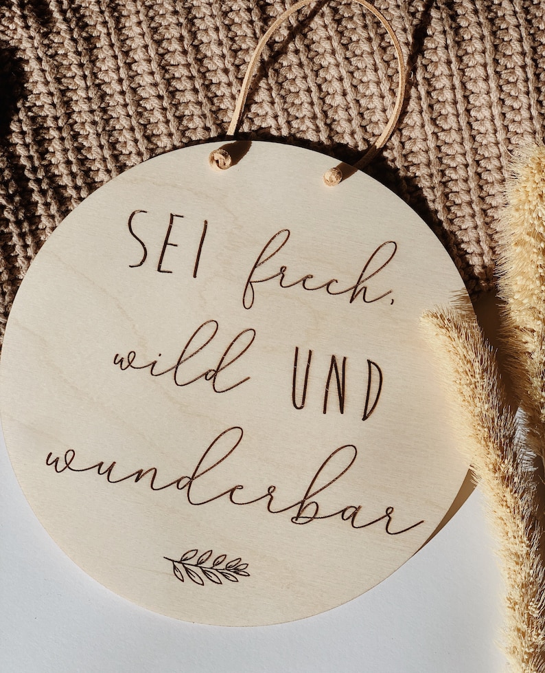 May include: A round wooden sign with the text "Sei frech, wild und wunderbar" (Be bold, wild and wonderful) in brown lettering. A small leaf design is below the text.