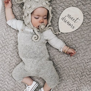 May include: A baby wearing a gray knitted hat with pom-poms and a gray knitted onesie is sleeping on a gray knitted blanket. The baby is wearing a pacifier and has a wooden sign with the text "sweet dreams" next to them.