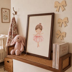 Può includere: Una stampa incorniciata in stile acquerello raffigurante una ballerina in tutù rosa, esposta su una mensola in legno con dettagli in paglia di Vienna. Accanto alla cornice si trovano un coniglio di peluche rosa e due libri intitolati Love You Tot Forever e Lana's World.