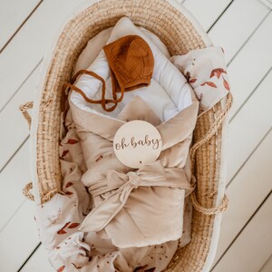 Pu&ograve; includere: Un fasciatoio per neonati in velluto beige con un berretto in tessuto marrone e un cartello in legno che dice "oh baby" in un cesto di vimini.