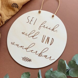 May include: A round wooden sign with a brown leather cord for hanging. The sign has the text "Sei frech, wild und wunderbar" (Be bold, wild and wonderful) in a cursive font.