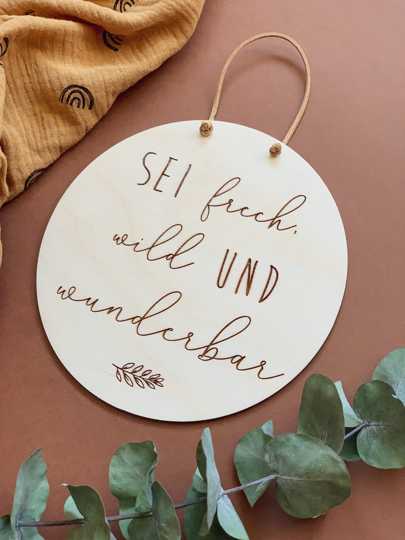 May include: A round wooden sign with a brown leather cord for hanging. The sign has the text "Sei frech, wild und wunderbar" (Be bold, wild and wonderful) in a cursive font.
