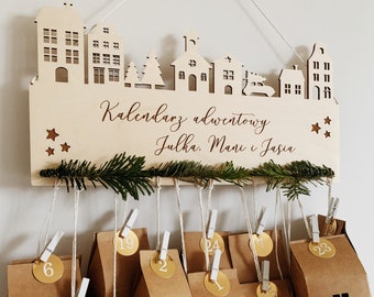 May include: Wooden Christmas advent calendar with a white background and a cutout design of houses and a Christmas tree. The calendar has 24 numbered paper bags hanging from it. The text "Kalendarz adwentowy Julka. Mani i Jasia" is written on the calendar.