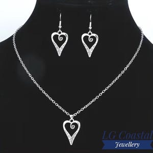 May include: A silver-toned jewellery set consisting of a necklace and matching drop earrings. Each piece features a heart-shaped pendant with an open centre and a small spiral detail at the top. The necklace includes a delicate link chain.