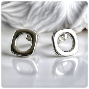 May include: A pair of silver square stud earrings with an open circle design. The earrings have a polished finish and are displayed on a white surface. The earrings are a modern jewellery design.
