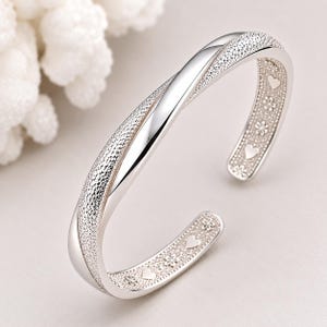 May include: A silver-toned open cuff bracelet featuring a twisted design with a textured, sparkling finish on the outer surface. The inner band is decorated with a repeating pattern of small engraved hearts and flowers along its entire length.