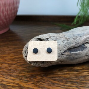 Puede incluir: Un par de pequeños pendientes de botón negros mate presentados en una tarjeta beige. Los pendientes redondos son de diseño sencillo, adecuados para el uso diario. La tarjeta descansa sobre madera flotante, con un fondo de madera.