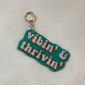 Puede incluir: Un llavero azul turquesa con un llavero dorado. El llavero tiene texto blanco que dice "Vibin' & Thrivvin'" en una fuente retro con contornos naranjas.