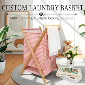 May include: A pink and white checkered laundry basket with a wooden frame. The basket is foldable and has a white towel hanging on the side. The text "CUSTOM LAUNDRY BASKET" and "Multiple Sizes/Multiple Colors/Foldable" is displayed above the basket.